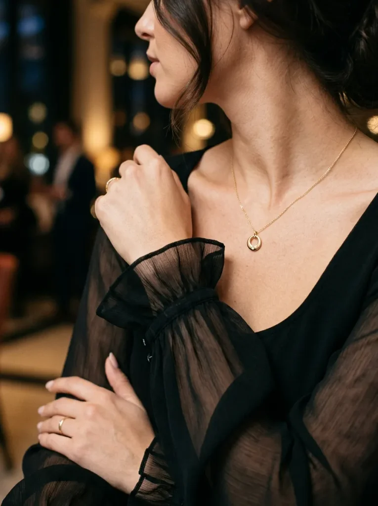 A close-up of a woman wearing an elegant black dress featuring a V-neckline and sheer chiffon long sleeves with ruffled cuffs. She is softly accessorized with a delicate gold circular pendant necklace and thin gold rings, with her hair pulled back loosely. The background shows softly blurred, warm ambient lighting typical of a formal evening event.