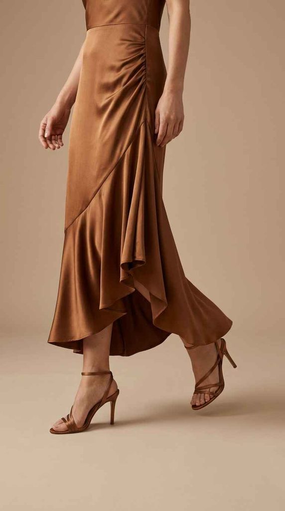 A photograph focusing on the lower half of a woman taking a step, highlighting the side waist gathering and the movement of the asymmetrical draped ruffled hem of a bronze satin midi dress flowing above bronze strappy heels.