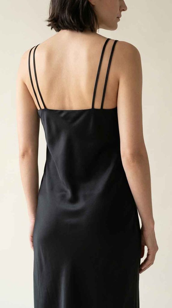 A photograph focusing on the back of a woman wearing a black satin slip dress, showing the sleek straight back neckline and double spaghetti straps.