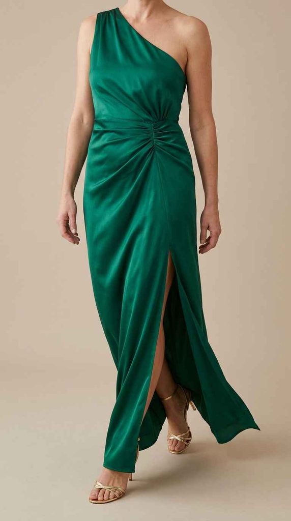 A photograph showing the lower half of a woman stepping forward in an emerald green satin maxi dress, emphasizing the gathered waist ruching, high side slit, and gold strappy heels.