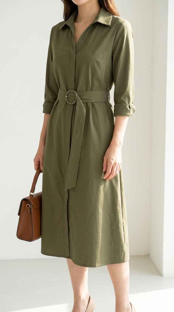A close-up photograph focusing on the upper body and waist of a woman wearing an olive green shirt dress with a round-buckle belt, carrying a brown top-handle handbag.